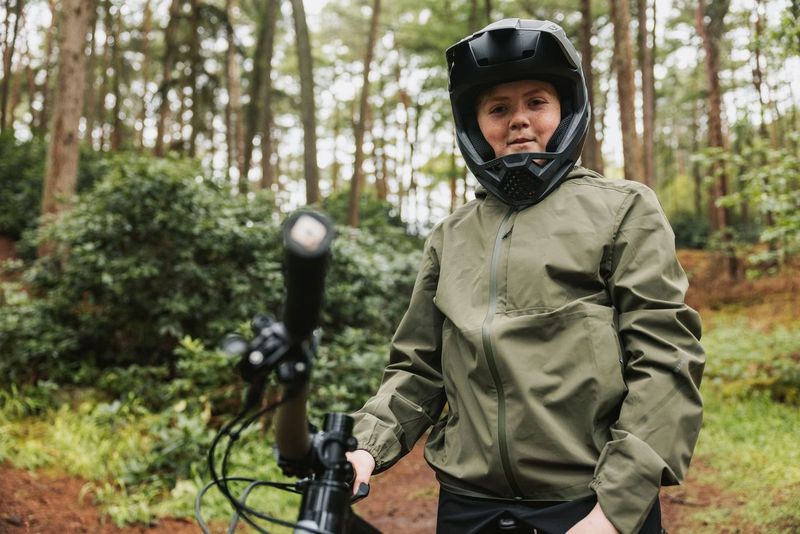 Person in green jacket and black helmet standing in a forest with a bicycle.