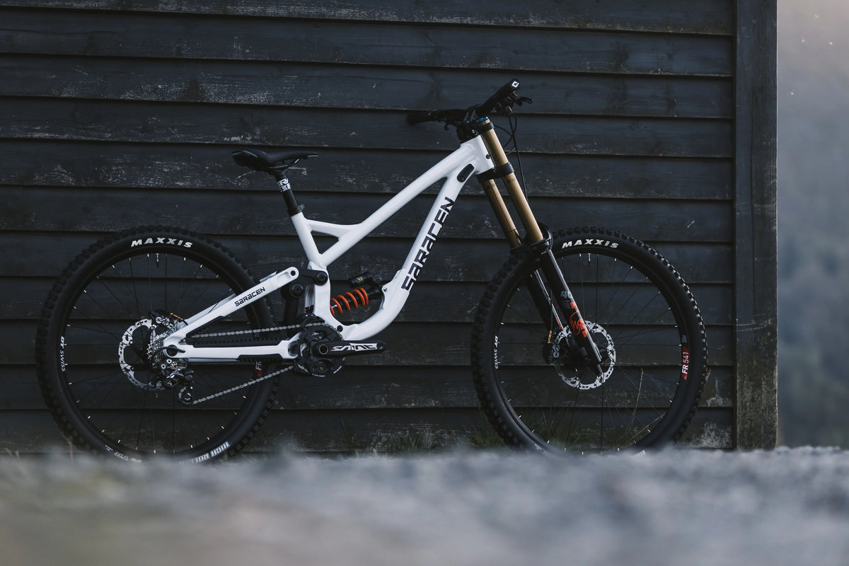 A white full-suspension mountain bike with black components and gold front fork accents, parked against a dark gray wooden wall.