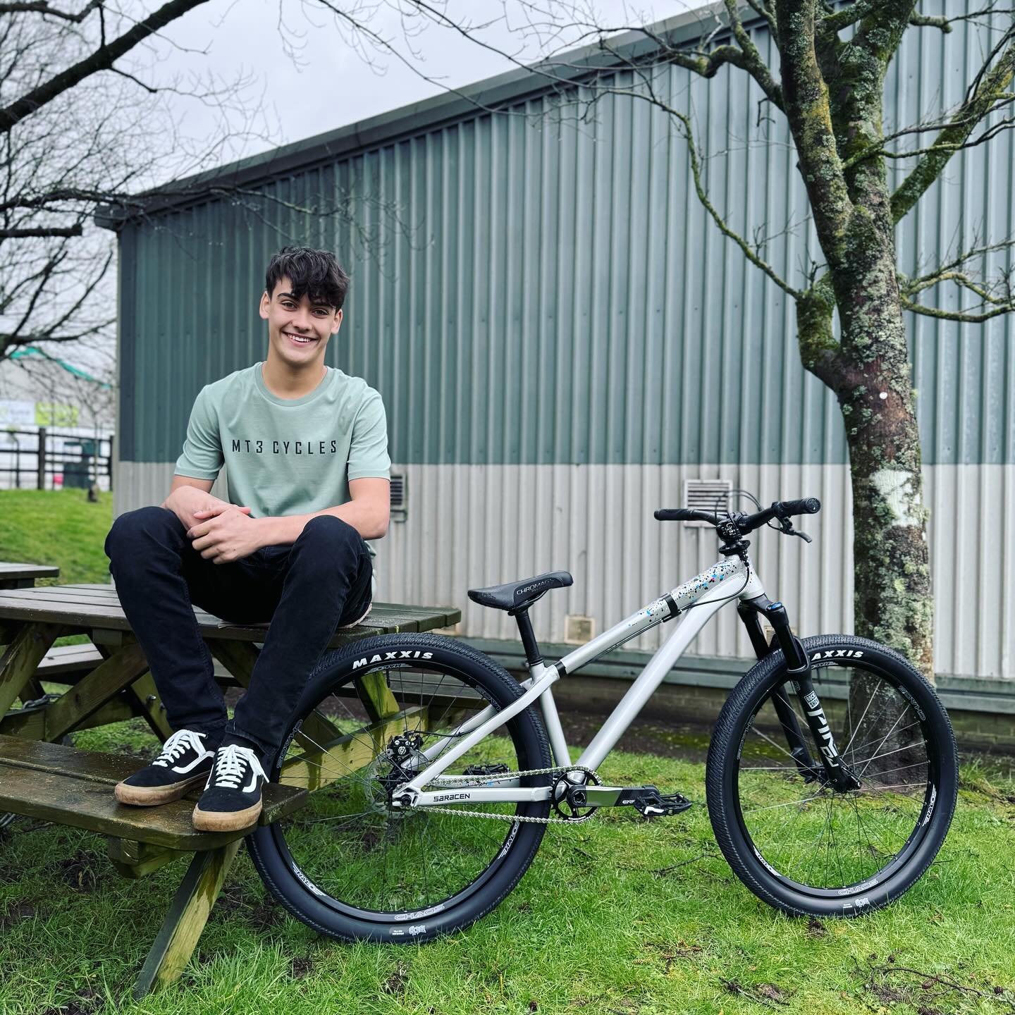Mountain biker sat on bench next to silver dirt jump bike, grass setting