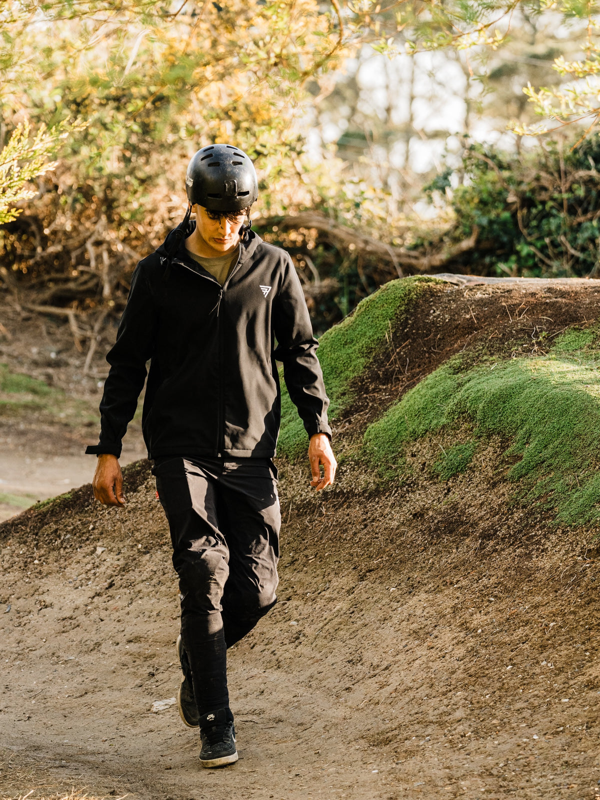 Person wearing a helmet and dark clothing walking on a dirt path with greenery in the background