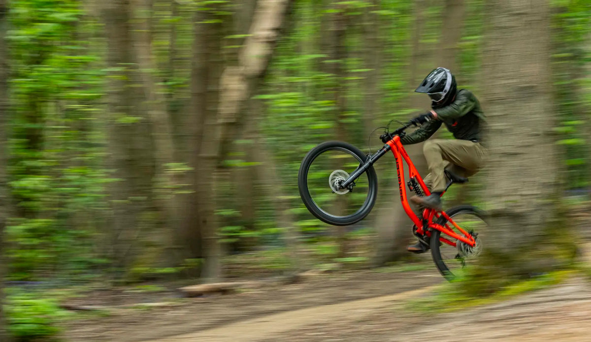 A mountain biker in a forest jumps a bright red full-suspension bike, captured in motion with a blurred green woodland background.