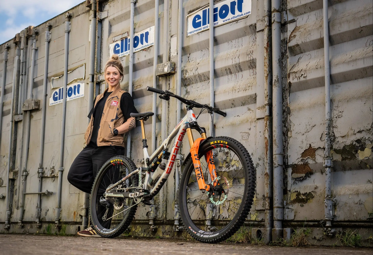 A woman stands next to a full-suspension mountain bike in front of a weathered shipping container. The bike has a silver frame with a bright orange front fork and black components.