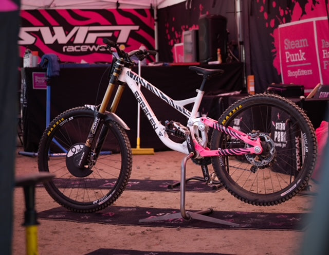 White and pink bicycle with 'Swift' branding on a stand at an event.
