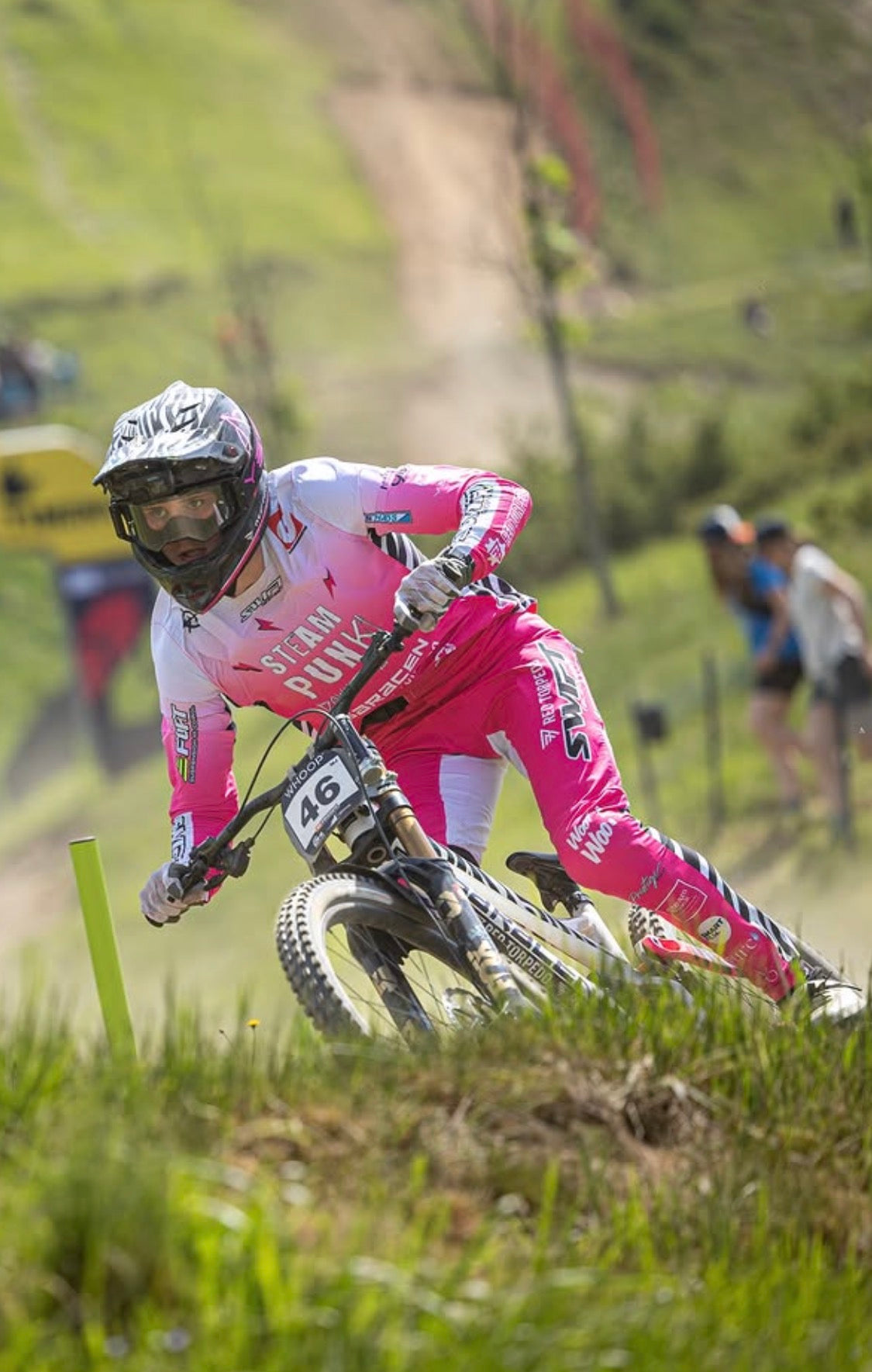 Biker in pink and white outfit riding a bike on a grassy trail with spectators in the background.