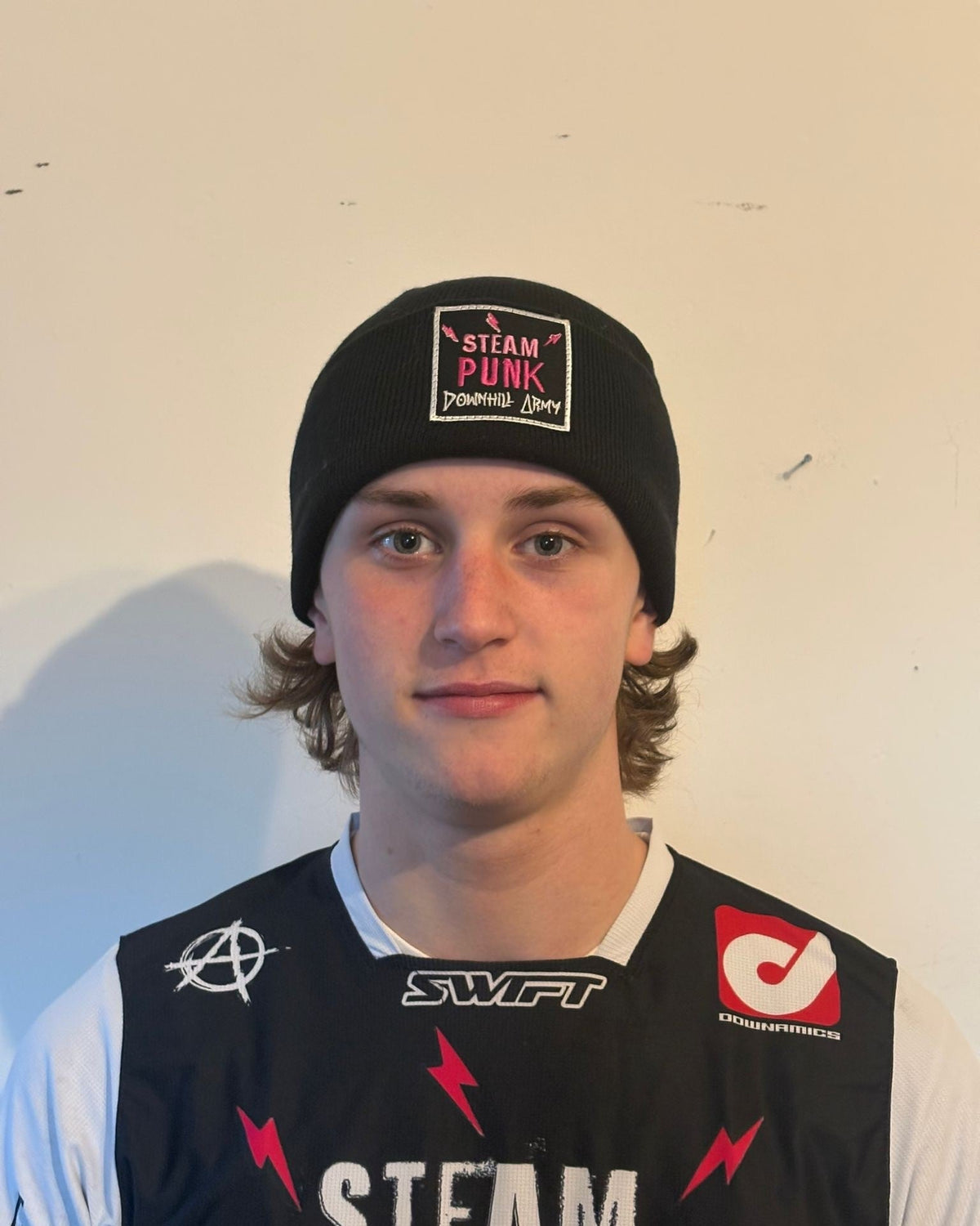 Person wearing a black beanie with 'Steam Punk' logo and a black sleeveless jacket with logos on a plain background