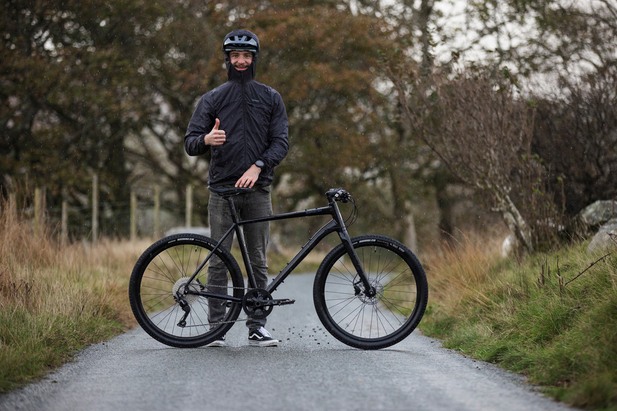 Person in black attire with a helmet holding a black bicycle on a rural road.