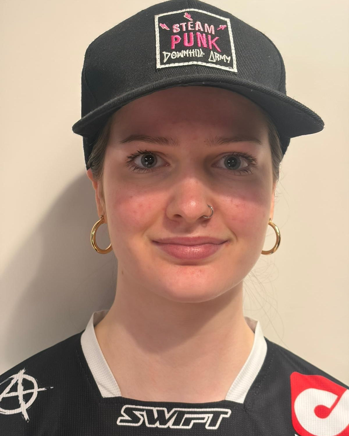 Person wearing a black cap with text and a black shirt with logos against a plain background