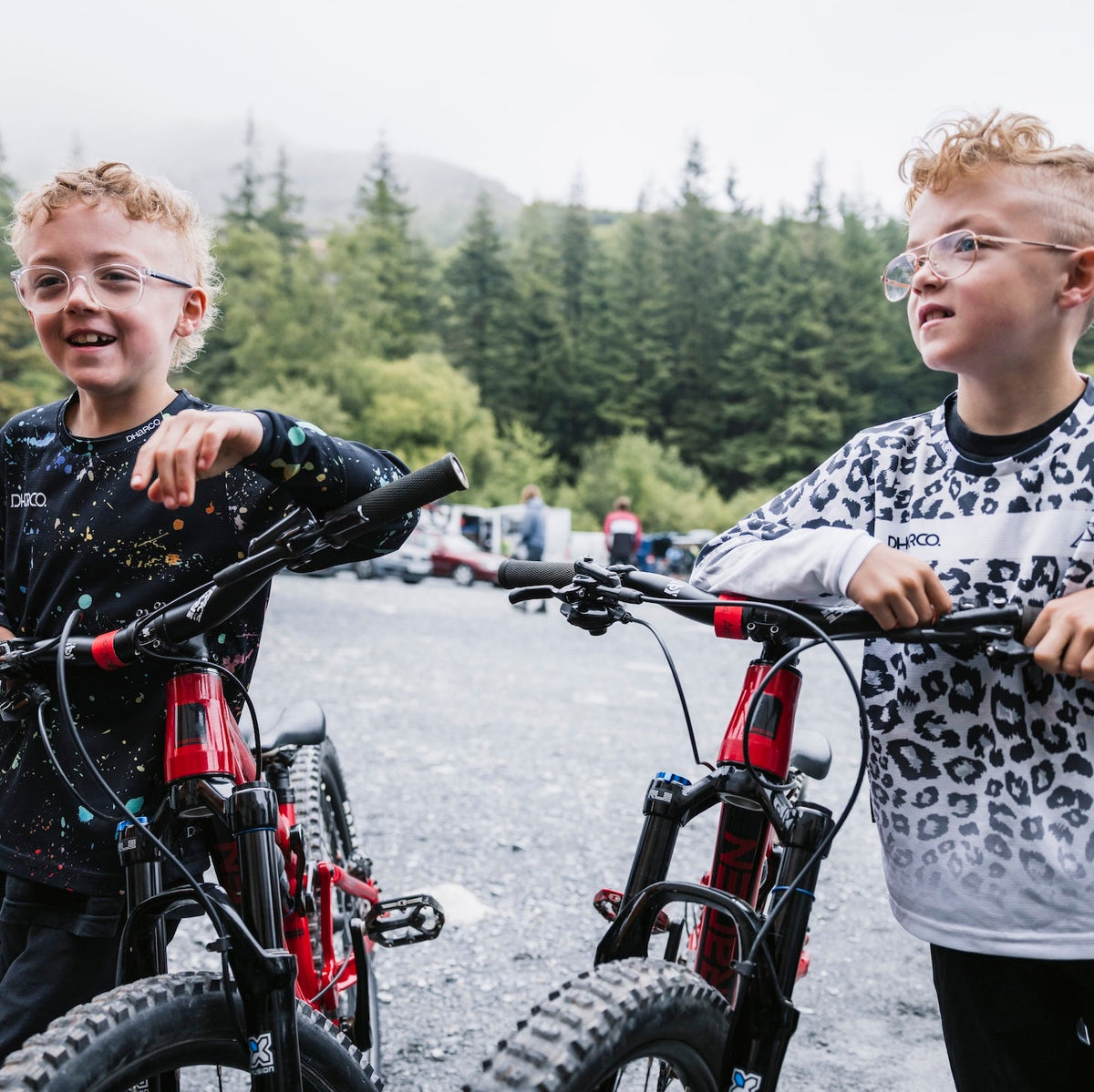 Two children with bicycles in an outdoor setting with trees and a sign in the background.