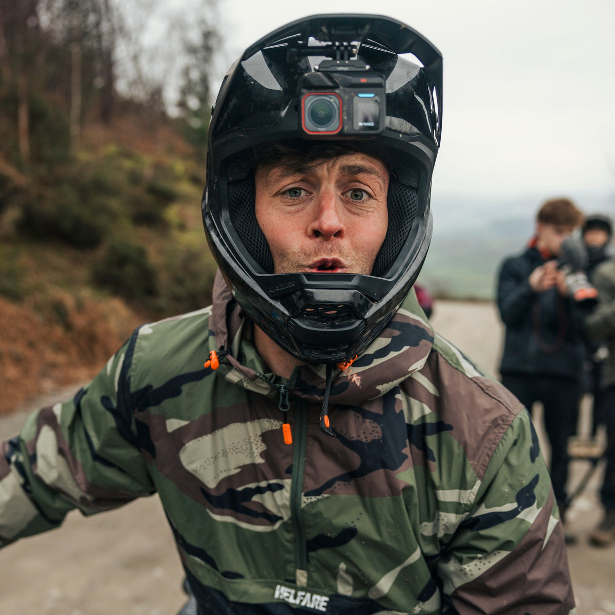 Person wearing a helmet with a camera, camouflage jacket, and gloves on a mountain bike.