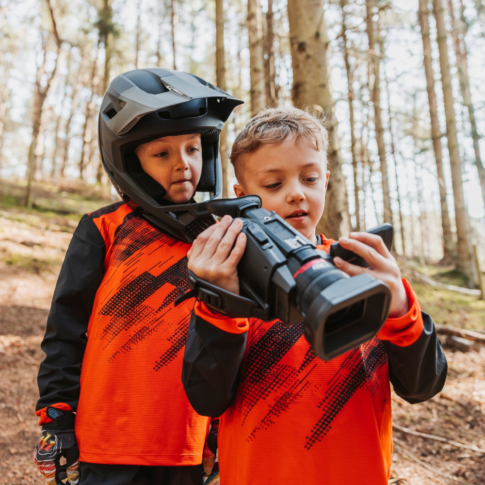Two children in orange and black outfits holding a toy camera in a forest setting.