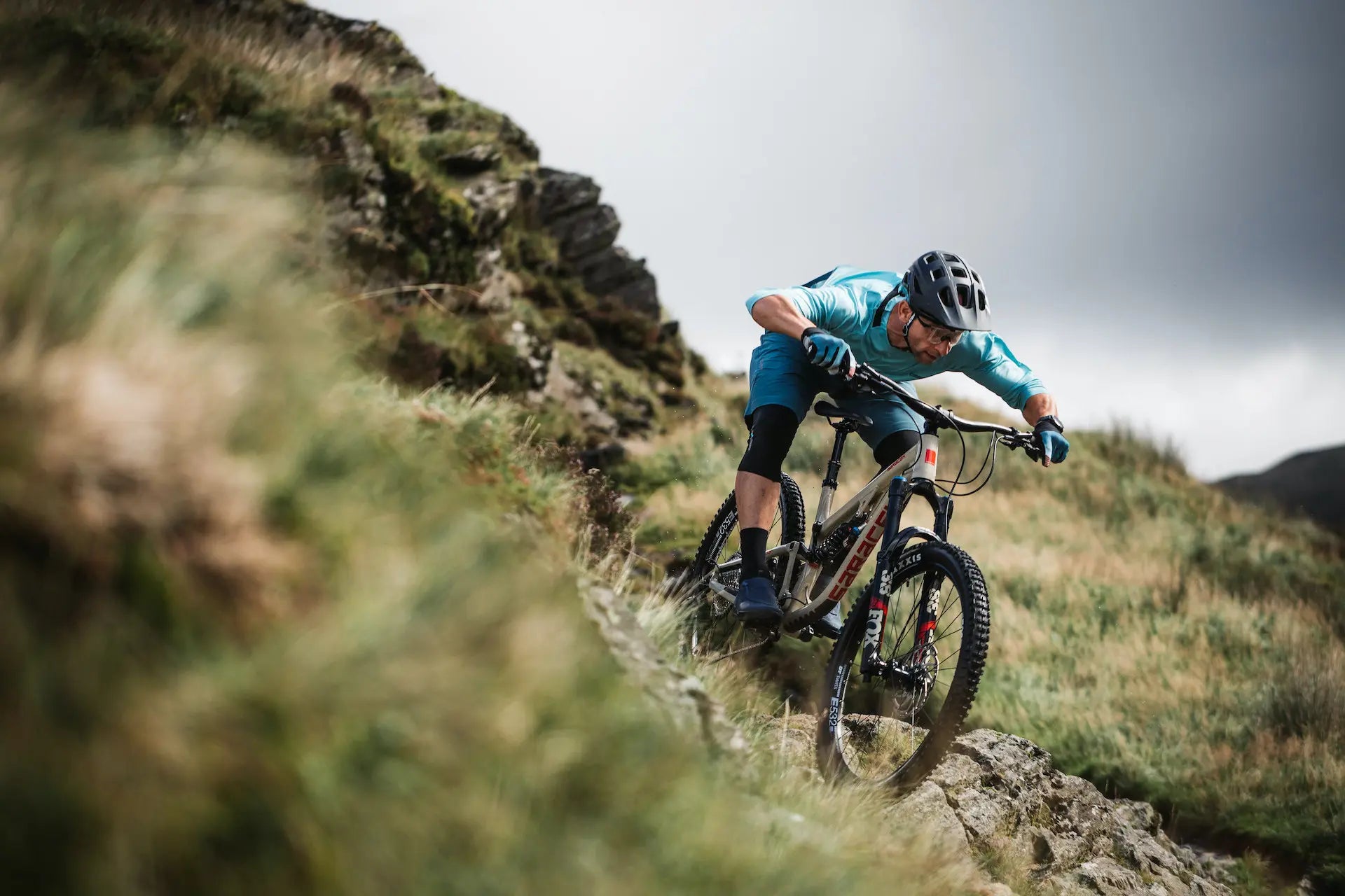 A mountain biker in a light blue jersey and black shorts leans sharply over a rocky trail on a grassy hillside under an overcast sky.