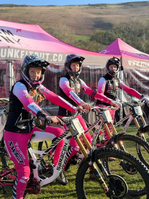Three individuals in pink racing suits with mountain bikes in front of a pink tent.