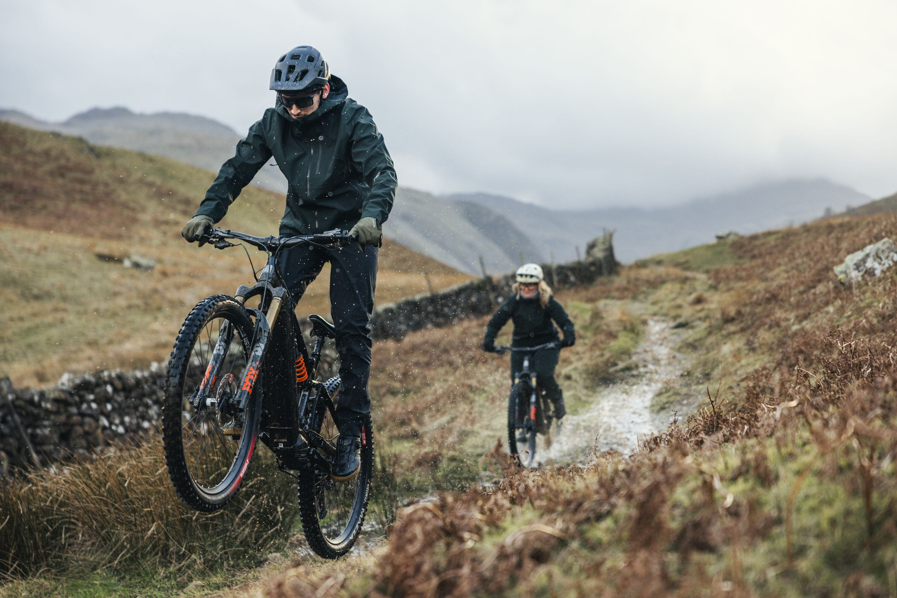 Two mountain bikers riding on a narrow dirt path through a rugged, grassy outdoor landscape under an overcast sky. The foreground rider wears a black helmet, goggles, and dark jacket and pants, while the second rider follows behind on a similar bike.