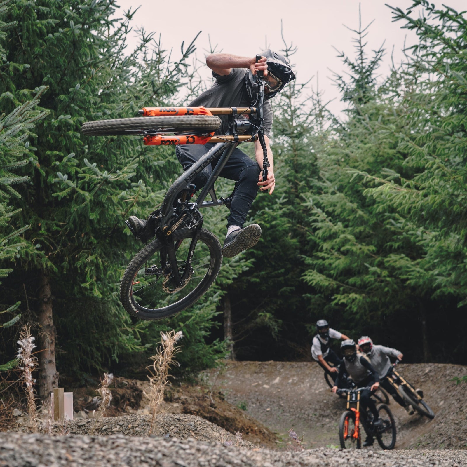 Person on a bicycle performing a jump in a forest setting with other cyclists in the background.