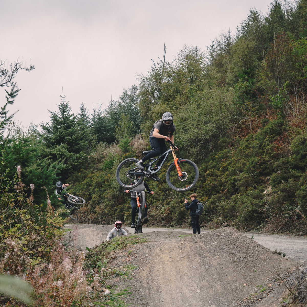 Person on a bicycle in mid-air over a dirt path with trees and other people in the background.